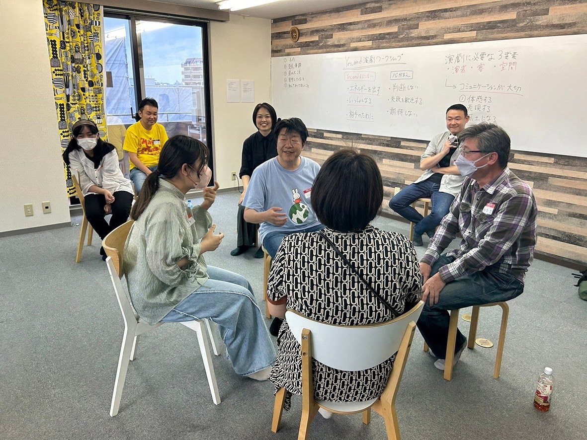 椅子で輪になり、笑顔で語り合う参加者たち。背後のホワイトボードにはYes,andの3原則が書かれている